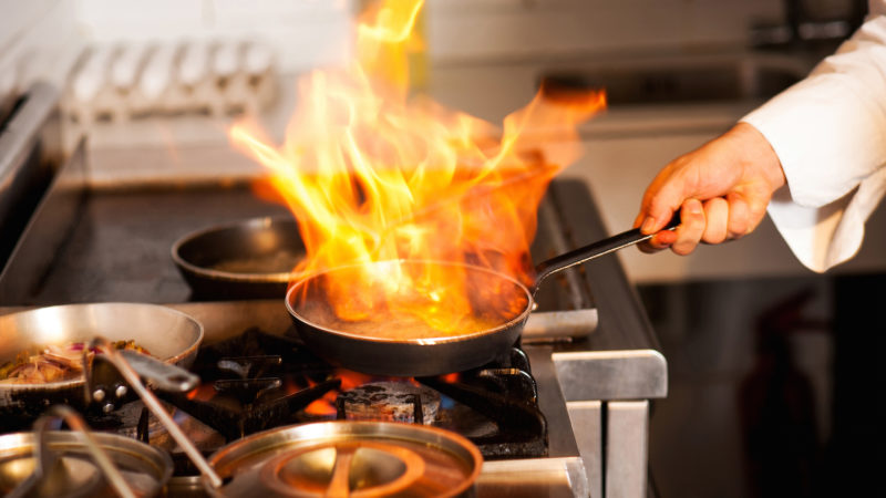 Chef cooking with flame in a frying pan on a kitchen stove.